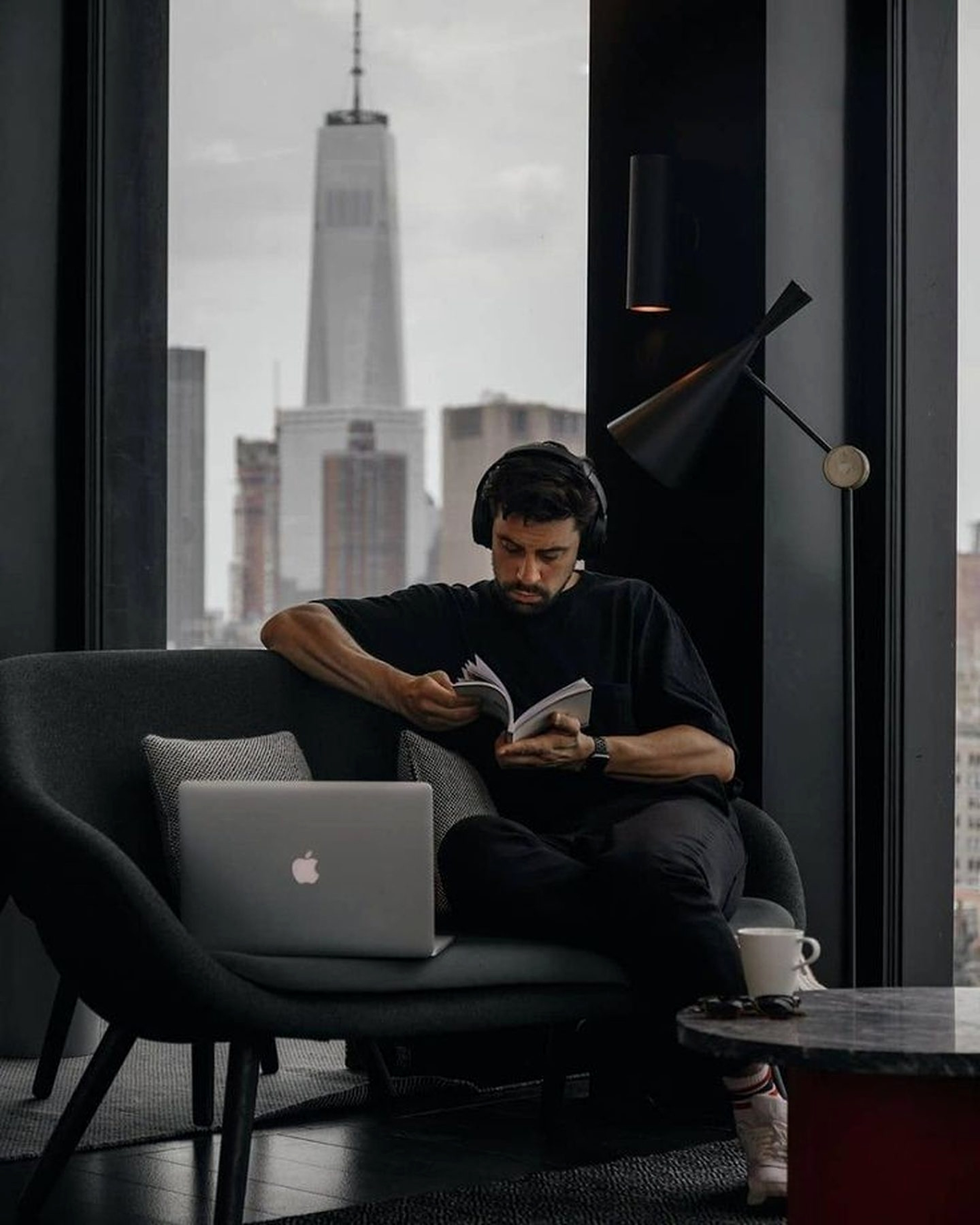A man reading at a low chair in a dark city apartment, a closed laptop on the table beside him, a coffee cooling, a quiet evening skyline through the window.