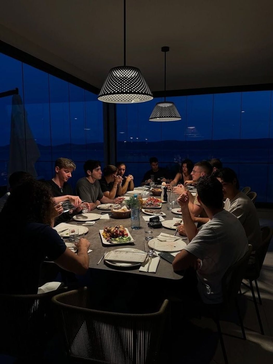 A long table of members at a private dinner, water visible through the windows beyond